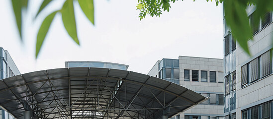 Auf dem Bild ist das Gebäude zu sehen, in dem das Bundesamt für Soziale Sicherung untergebracht ist. Man sieht von Weitem den Eingangsbereich mit einem Unterstand und oben im Bild ragen grüne Blätter von einem Baum ins Bild hinein.